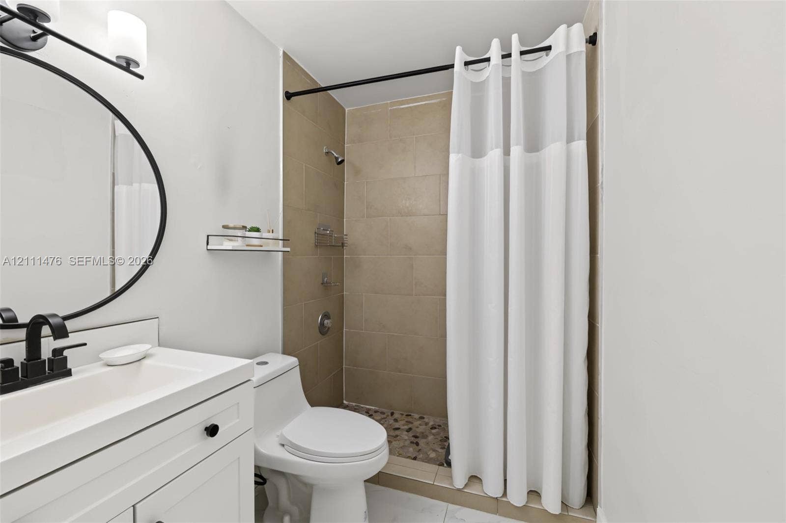 White vanity with integrated rectangular sink and matte black faucet, round wall-mounted mirror, matte black vanity lighting, shower enclosure with neutral-toned wall tiles, and stone-tiled shower flooring