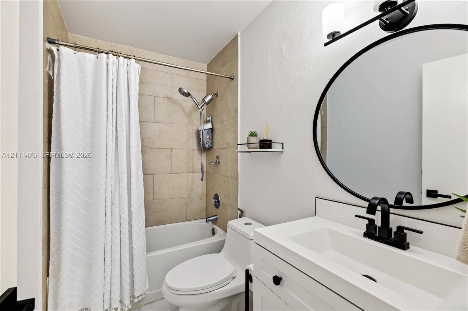 Bathroom featuring a white vanity with an integrated basin, matte black widespread faucet, circular mirror with a black frame, wall-mounted vanity lighting, and a bathtub with beige tiled surround