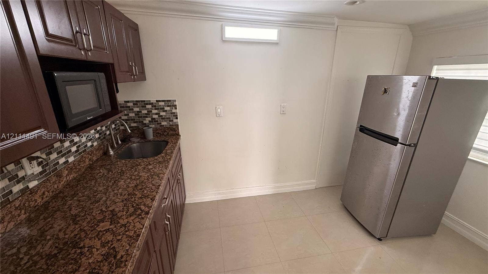 Kitchen area featuring dark-tone cabinetry, speckled stone-finish countertops, multi-toned glass tile backsplash, stainless steel sink with high-arc faucet, and tiled flooring