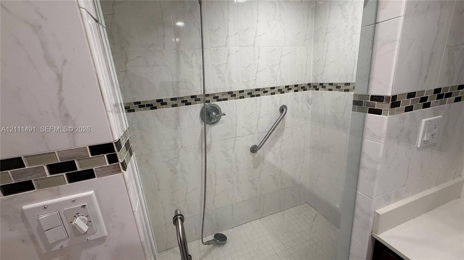Walk-in shower enclosure featuring floor-to-ceiling large-format stone-patterned wall tile, decorative mosaic tile accents, a handheld shower fixture, and wall-mounted support bars