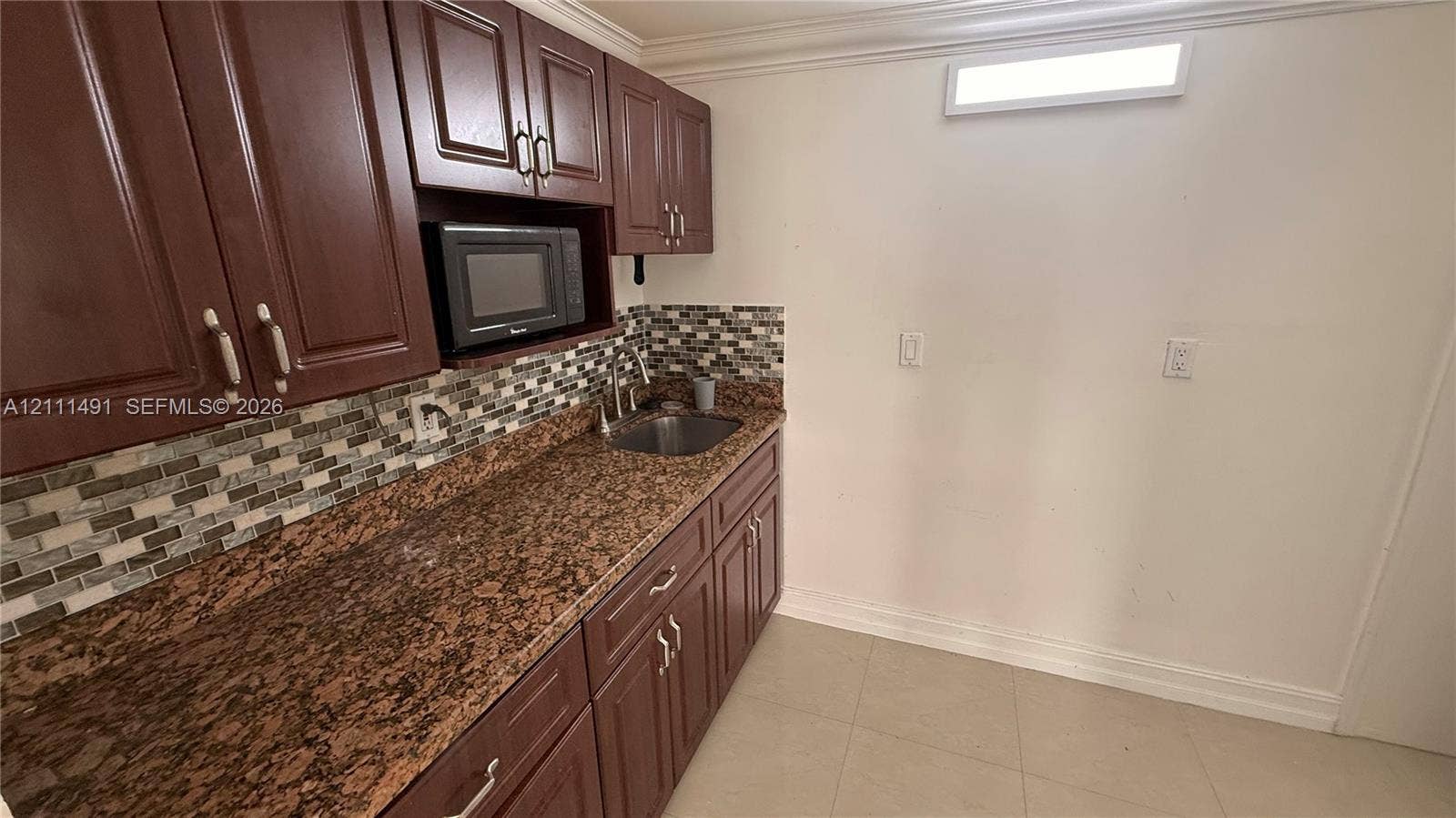 Dark wood-finish cabinetry, stone-texture countertop, mosaic tile backsplash, integrated stainless steel sink, and built-in microwave