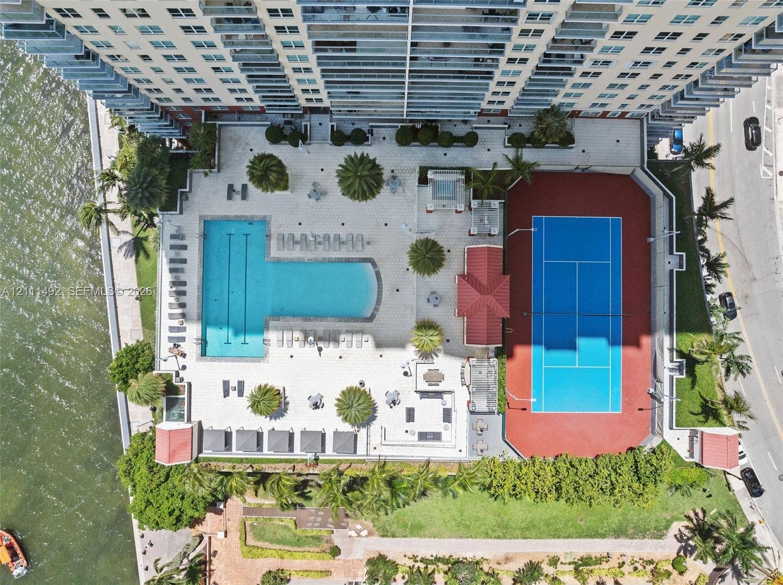 Aerial perspective of property amenities featuring a swimming pool, tennis court, tiled pool deck, palm trees, and landscaped grounds