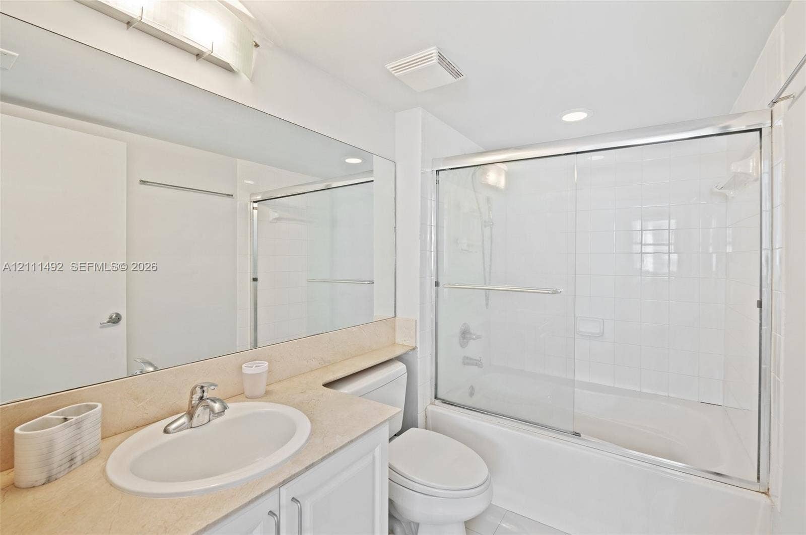 Full-width vanity with solid-surface countertop and integrated oval sink, expansive wall-mounted mirror, white cabinetry, and shower-tub combination featuring a sliding glass enclosure and full-height tiled surround