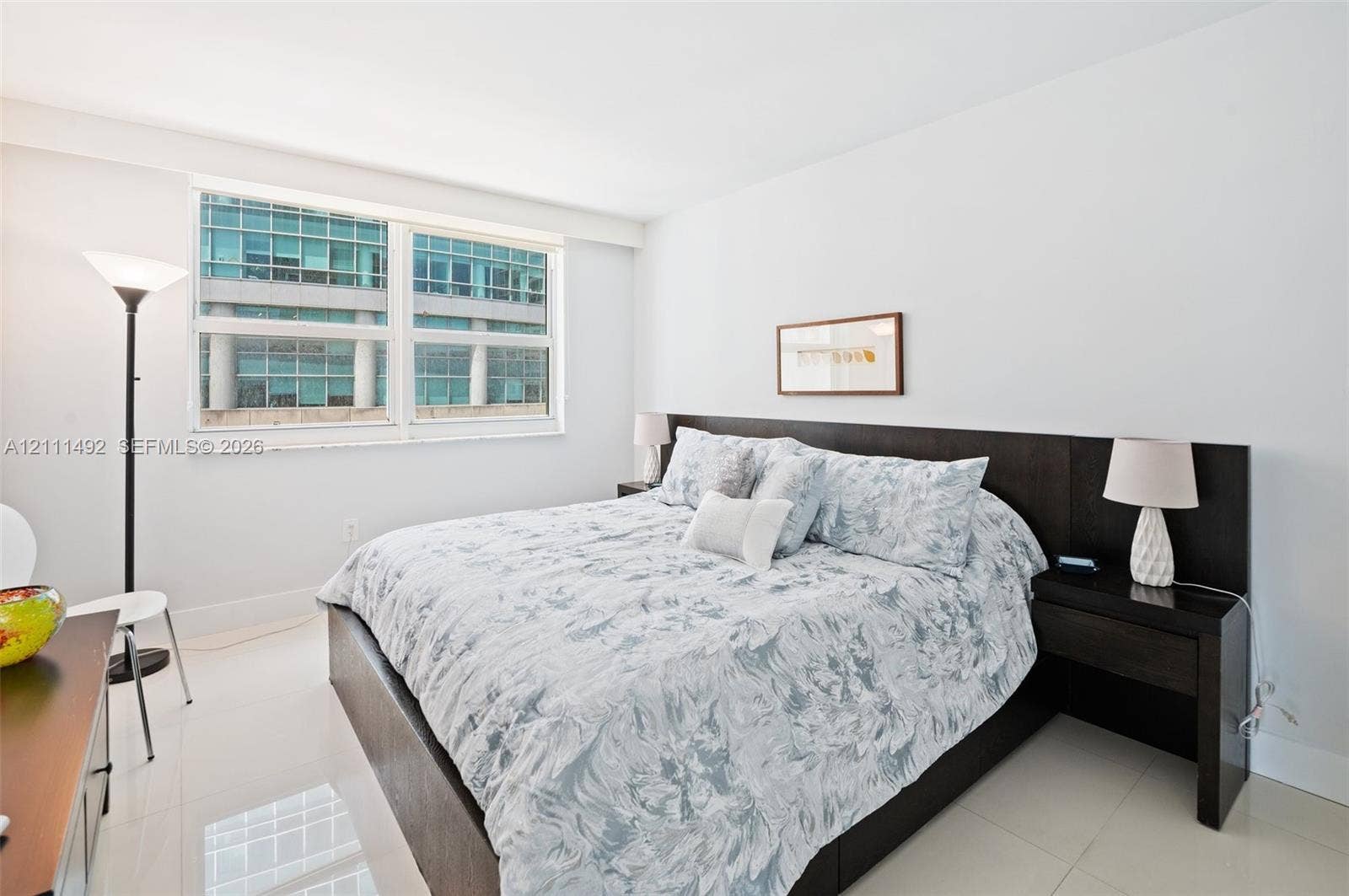 Large window with white frame, integrated dark wood headboard with built-in nightstands, and light-toned tile flooring