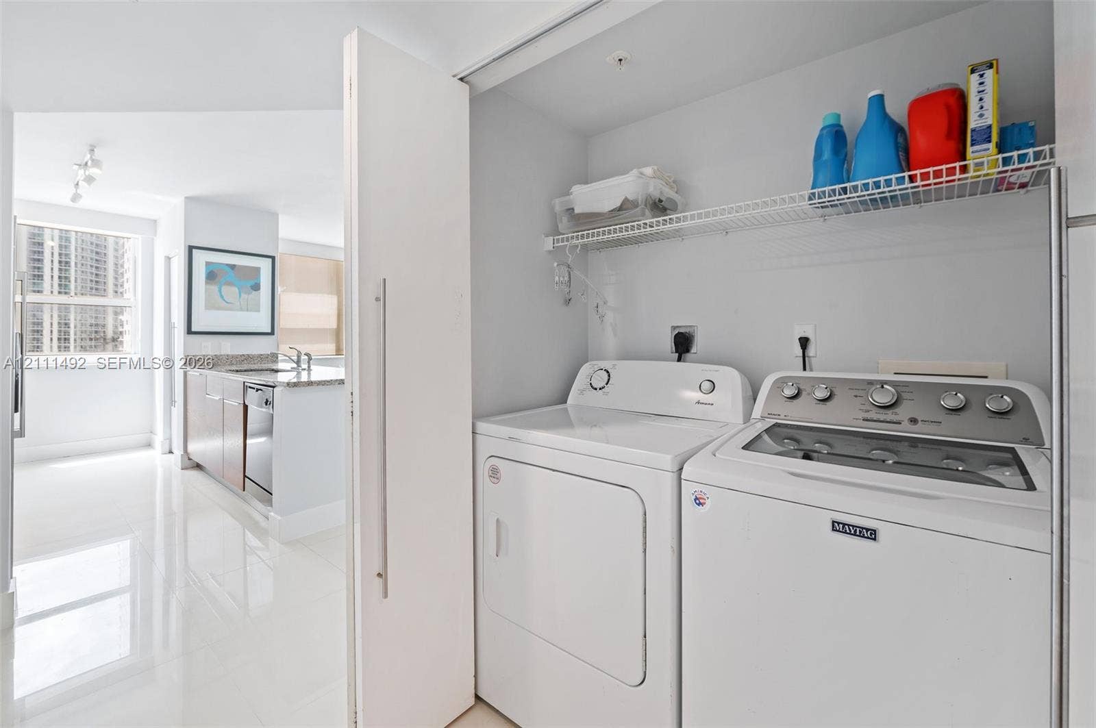 Laundry closet featuring a wire shelving unit and stacked washer and dryer, polished tile flooring, and a sleek kitchen space with stone-surfaced countertops and stainless steel appliances