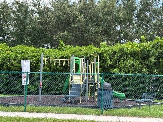 3210 SE 6th Street, Homestead, Homestead, FL 33033 Playground area featuring a multi-component play structure with a slide and climbing features, enclosed by green chain-link fencing, surrounded by mature perimeter hedging, and incorporating a permanent waste receptacle and stationary bench