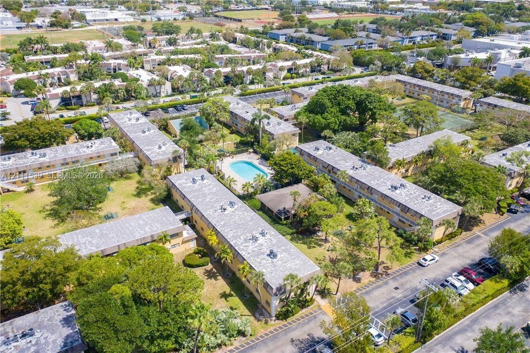 Aerial perspective of the residential complex featuring multi-story structures, an outdoor swimming pool, paved parking areas, and mature landscaping