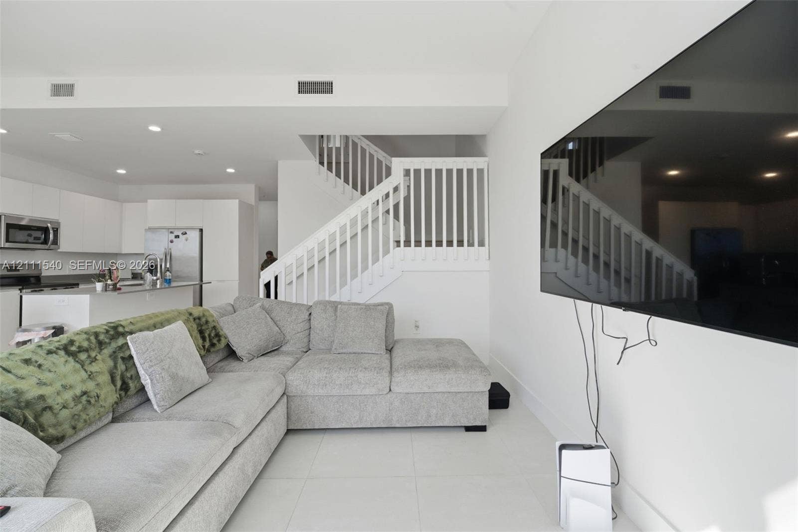Open-concept interior featuring light-toned tile flooring, recessed ceiling lighting, a staircase with white balusters, and a kitchen equipped with white cabinetry