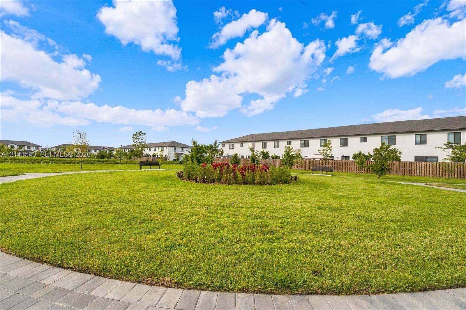 Expansive communal green space featuring a manicured lawn, established landscaping, paved walking paths, and fixed outdoor seating