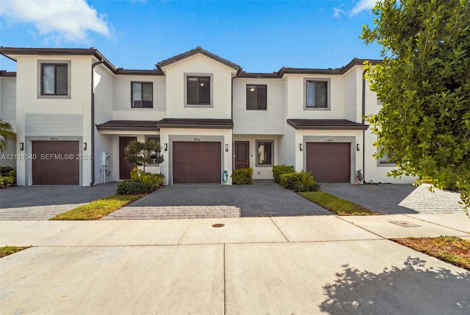 Multi-story townhouse exterior featuring a light-toned stucco facade, dark-finish single-bay garages, stone-paved driveways, and dark-trimmed windows