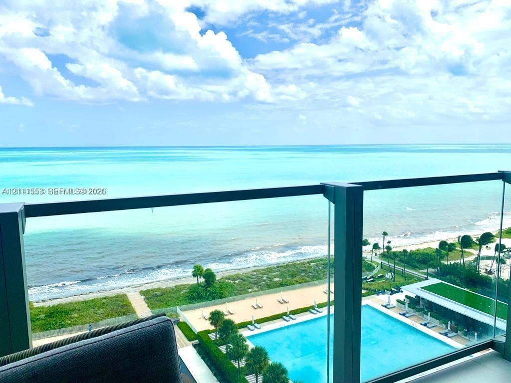 Private balcony featuring a glass-panel railing, overlooking a rectangular swimming pool, manicured grounds, and the ocean