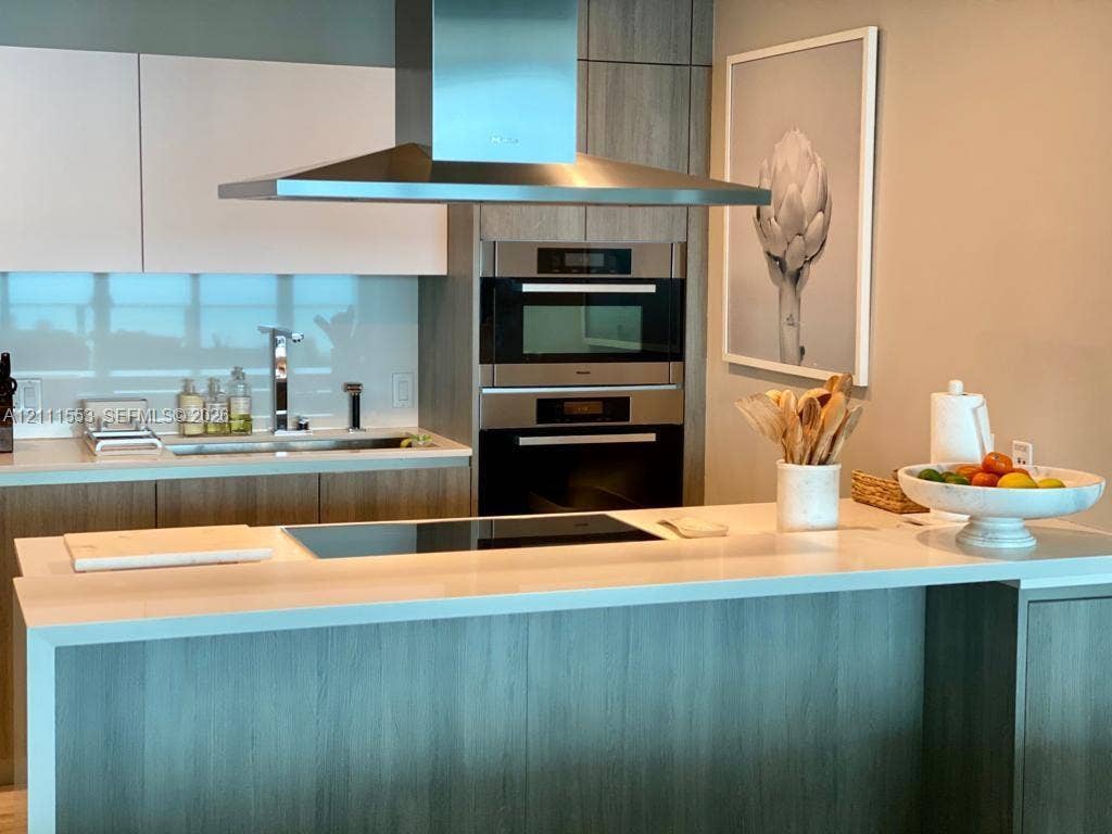 Modern kitchen featuring a waterfall-edge countertop, stainless steel chimney-style range hood, stacked built-in wall ovens, wood-finish cabinetry, and a glass tile backsplash
