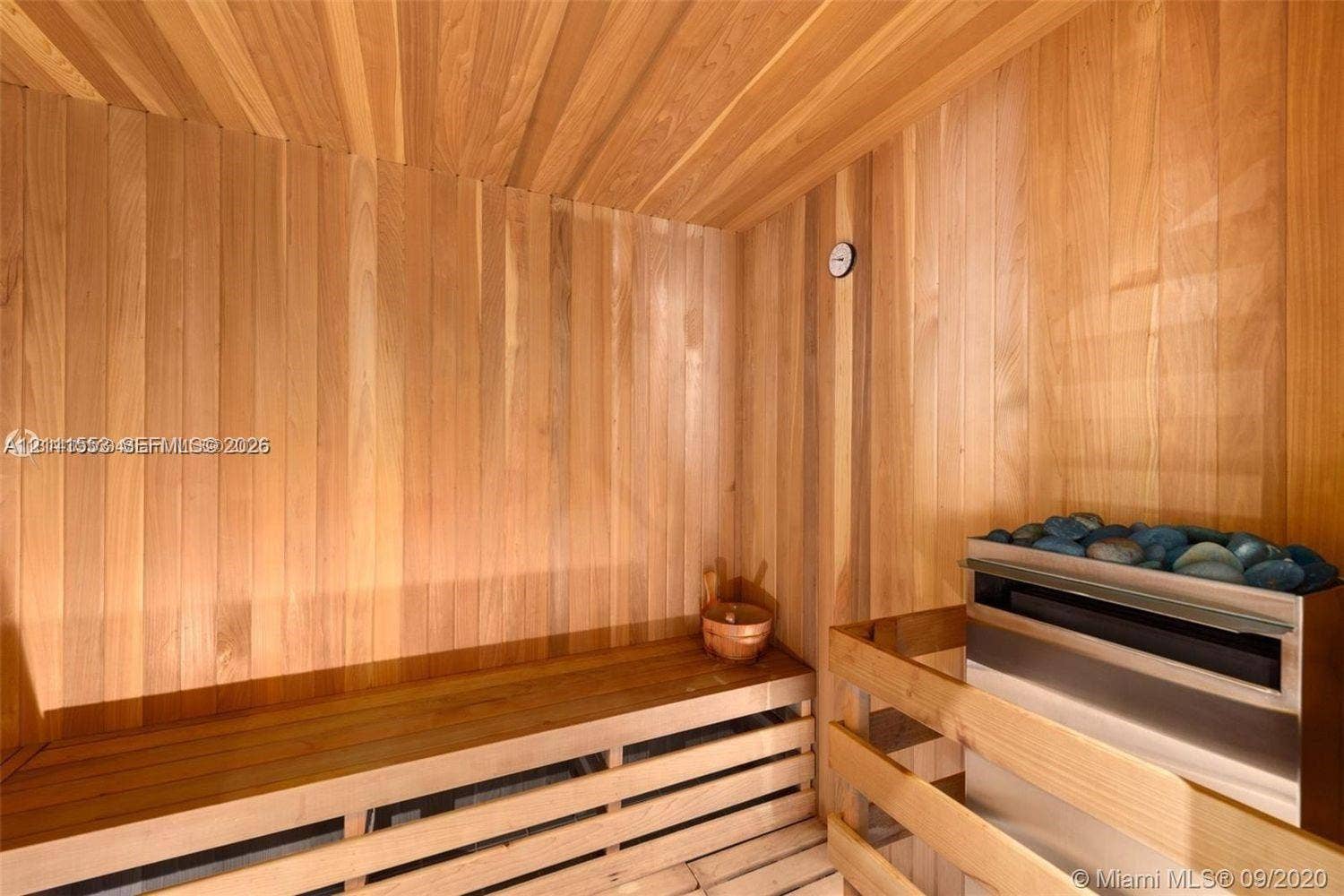 Sauna featuring wood-paneled walls and ceiling, slatted wood bench seating, and a stainless steel heater with stone fill