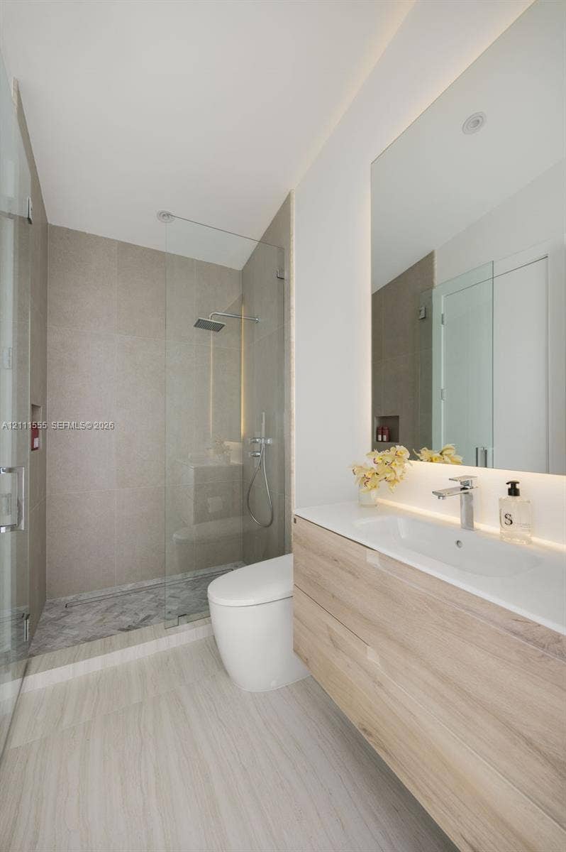 8800 SW 118th Street, Miami, Miami, FL 33176 Floating wood-finish vanity with an integrated basin, frameless glass walk-in shower featuring a rainfall showerhead, neutral stone-finish tiling, and an expansive wall-mounted mirror with integrated backlighting