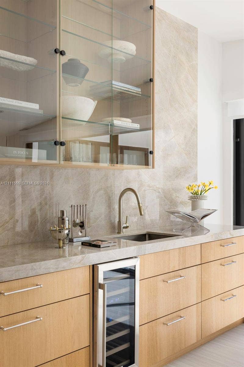 8800 SW 118th Street, Miami, Miami, FL 33176 Wet bar featuring light-toned wood cabinetry, stone slab backsplash and countertop, under-counter wine refrigerator, and upper glass-front cabinetry with interior shelving