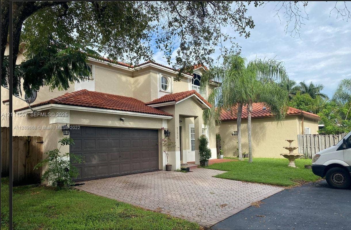 21514 SW 90th Avenue, Cutler Bay, FL 33189 Stucco exterior residence featuring a multi-gabled tile roof, paver driveway, attached two-car garage, and tiered garden fountain