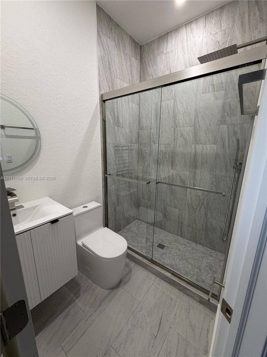 Bathroom featuring a walk-in shower with sliding glass doors, floor-to-ceiling stone-look wall tile, a vertical-ribbed vanity cabinet with a composite countertop, and large-format porcelain tile flooring
