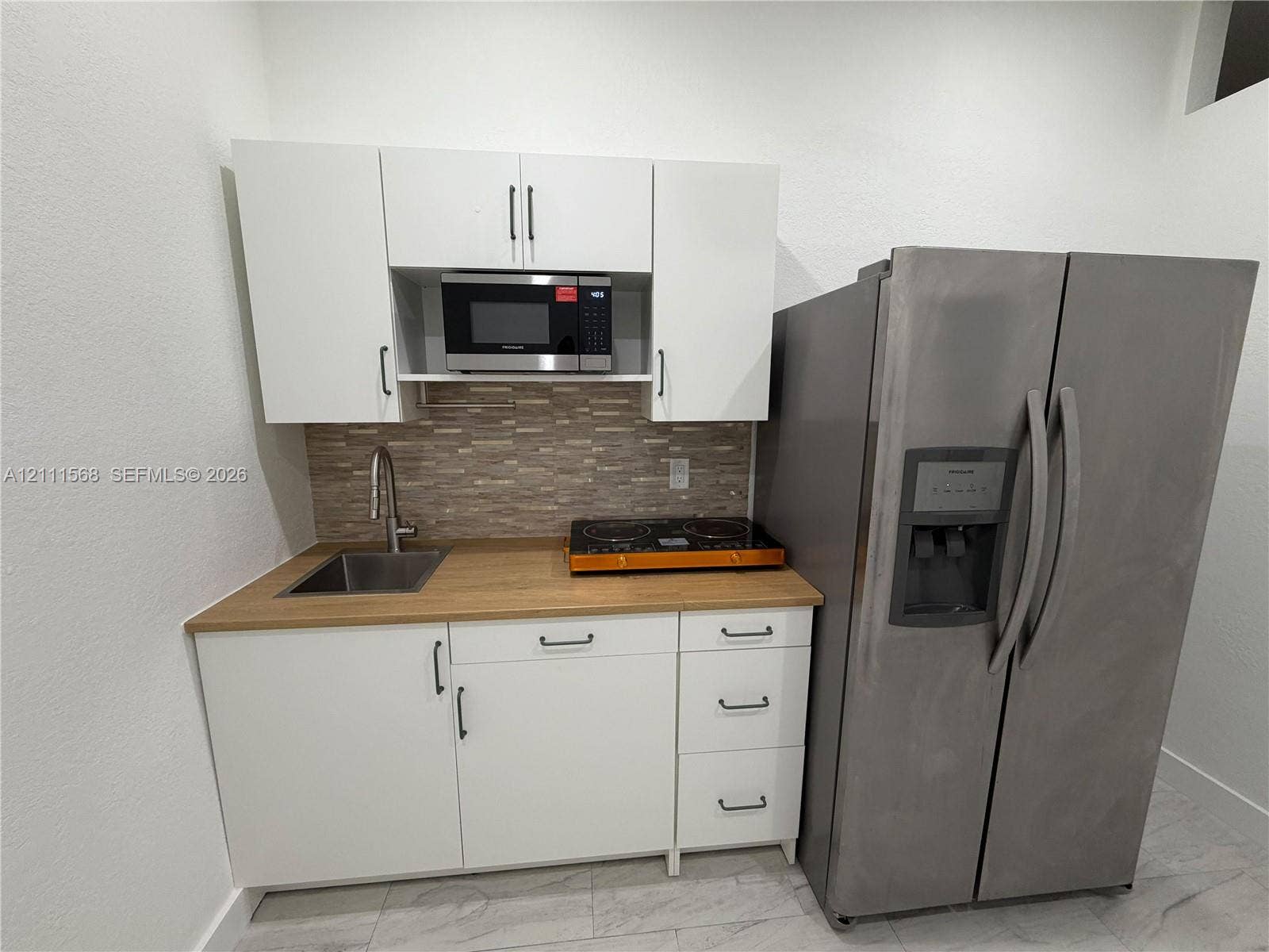 Kitchenette featuring white cabinetry, light wood-finish countertop, stainless steel sink with high-arc faucet, tile backsplash, and full-size stainless steel refrigerator