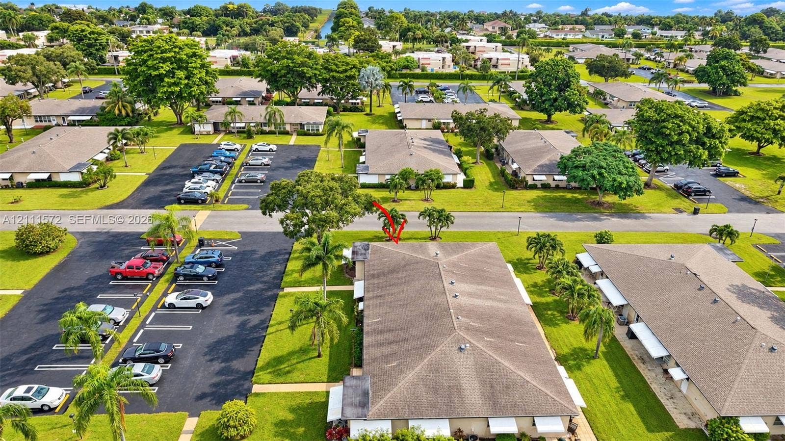 Aerial perspective of a well-maintained residential complex featuring low-rise structures with shingle roofing, paved surface parking, and extensive green space with mature palm trees