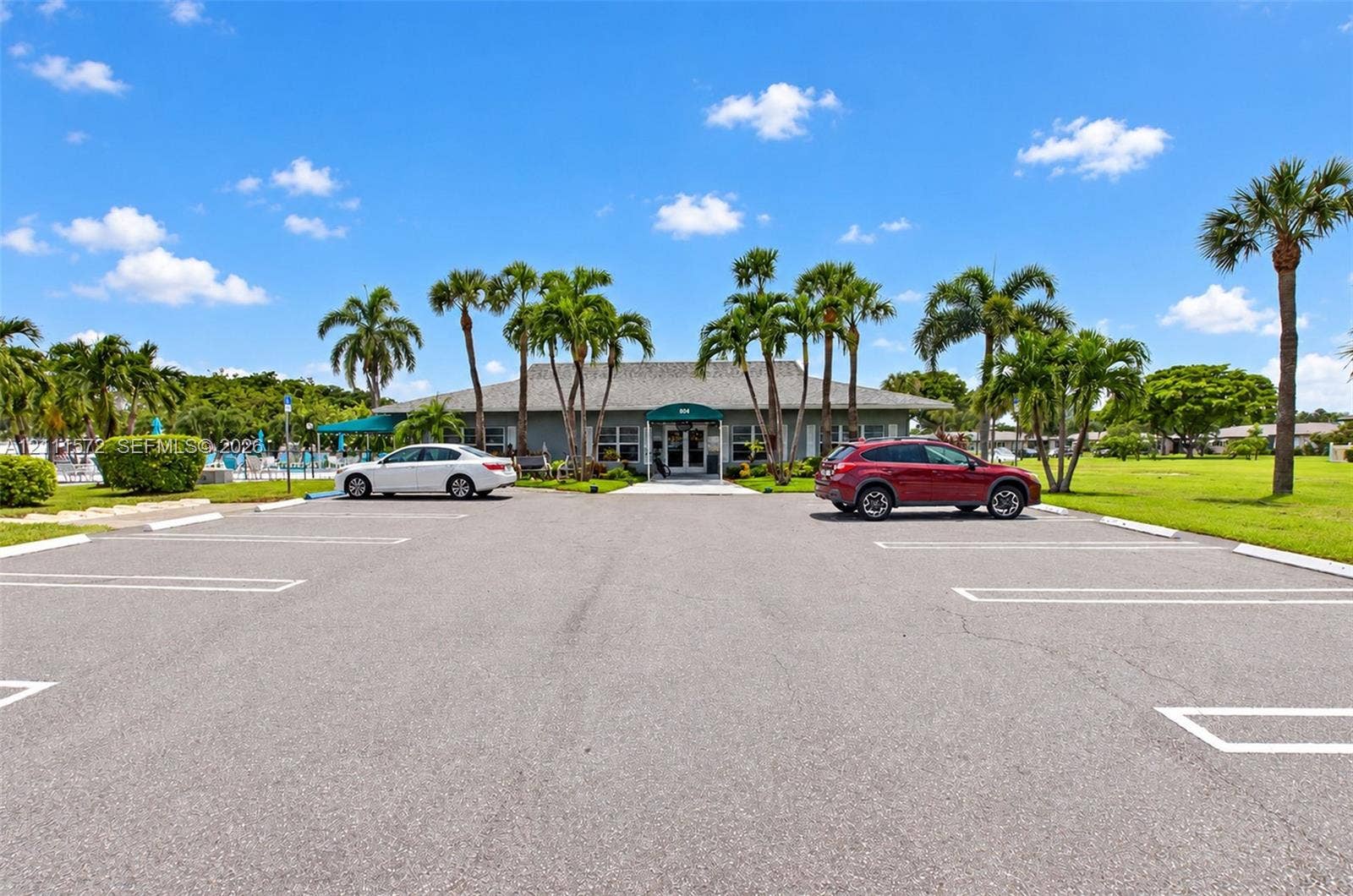 Paved surface parking lot with marked stalls, mature palm trees, and single-story building featuring a central covered entryway