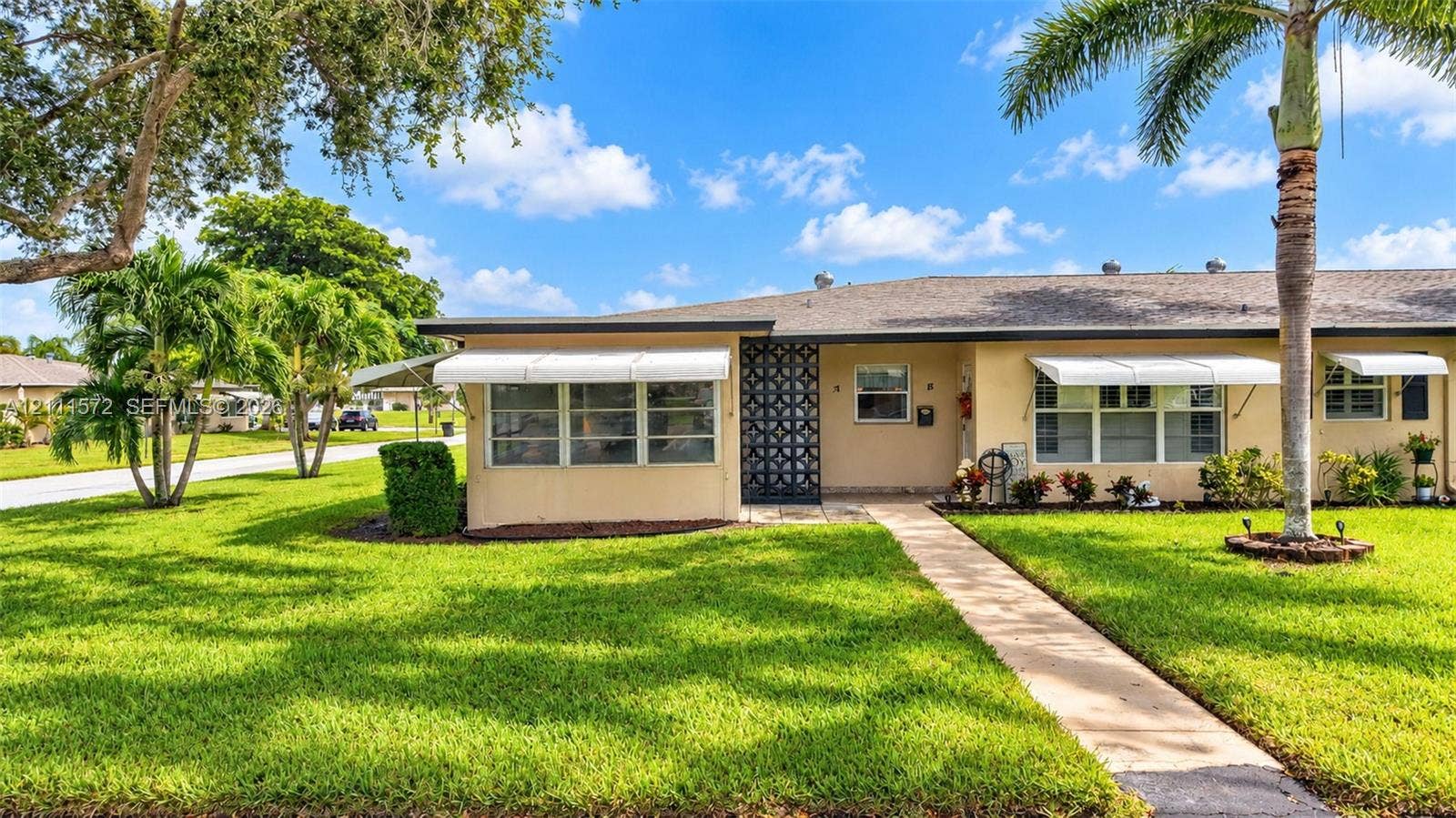 Single-story exterior villa featuring a light-toned stucco facade, decorative geometric breeze block entryway screen, metal window awnings, and a landscaped lawn with palm trees
