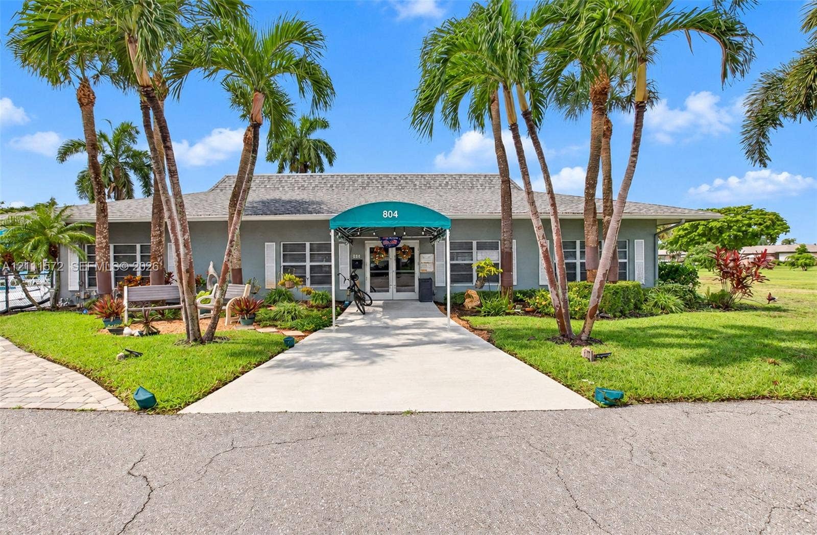 Exterior facade featuring a covered entryway with a teal fabric awning, concrete walkway, palm tree landscaping, and shingle roofing