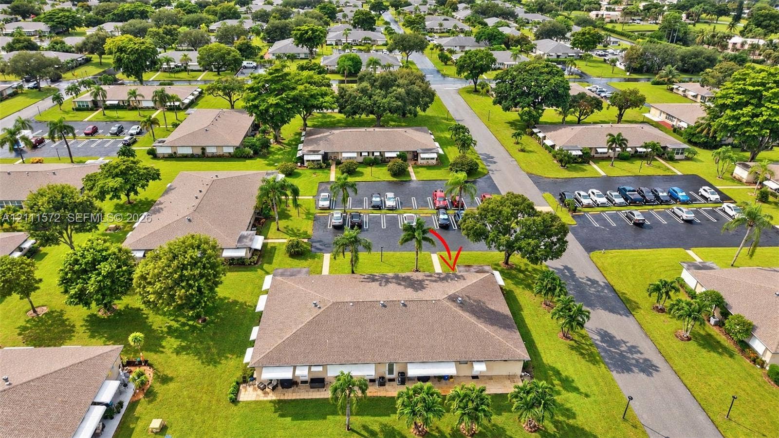 Aerial perspective of a single-story multi-unit structure featuring a shingled roof, paved parking areas, expansive green space, and mature landscaping