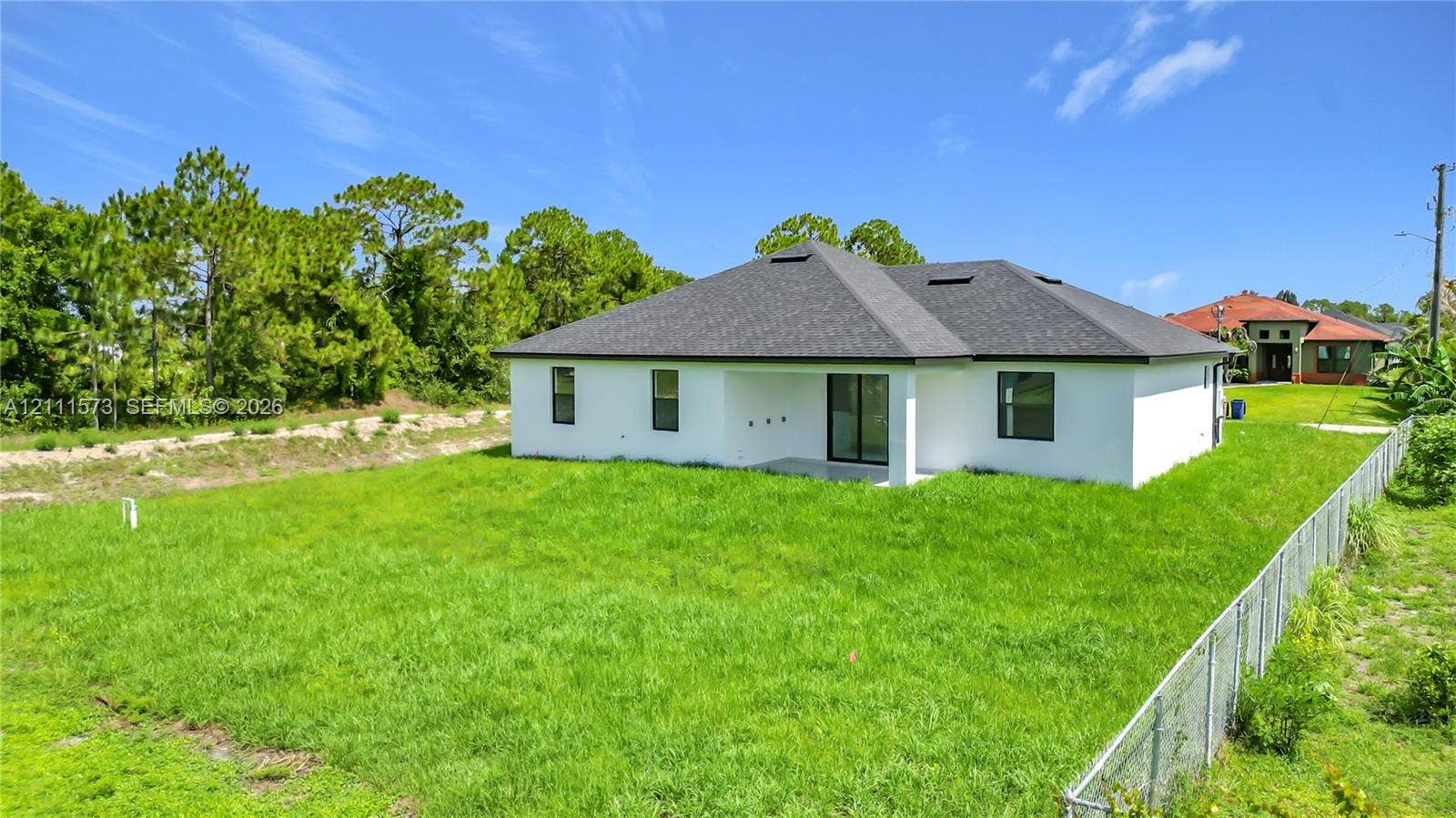 3813 SW 26th Street, Lehigh Acres, FL 33976 Exterior rear elevation featuring a white stucco facade, a dark shingled roof, a covered concrete patio, and a fenced-in grassy lot