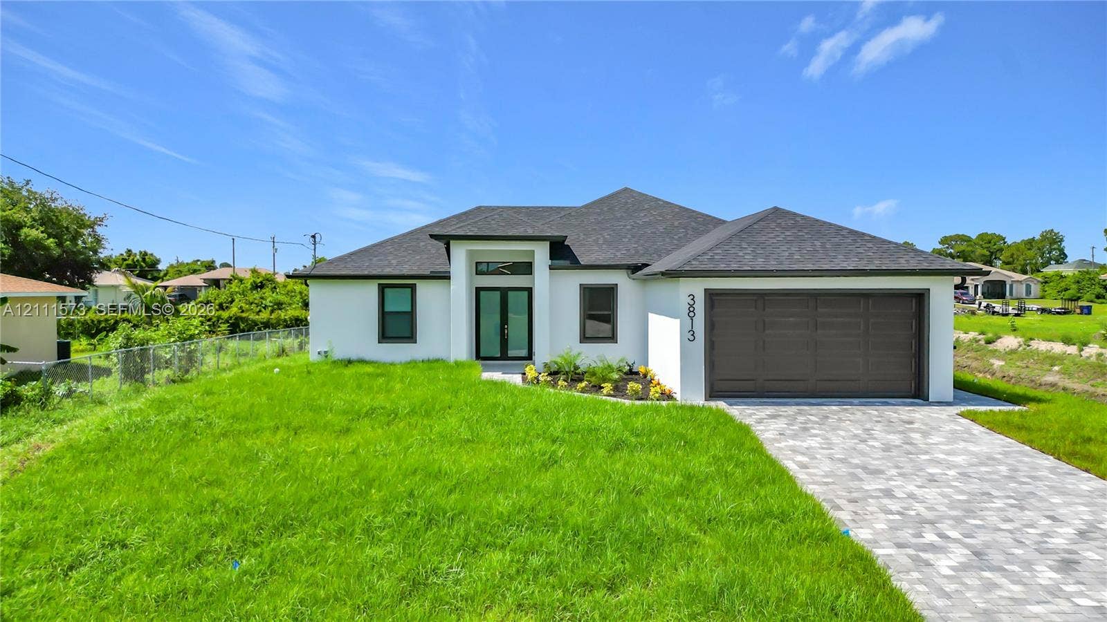 3813 SW 26th Street, Lehigh Acres, FL 33976 Single-story exterior featuring white stucco siding, a grey shingle roof, double-entry doors with transom window, and a paver driveway leading to an attached two-car garage