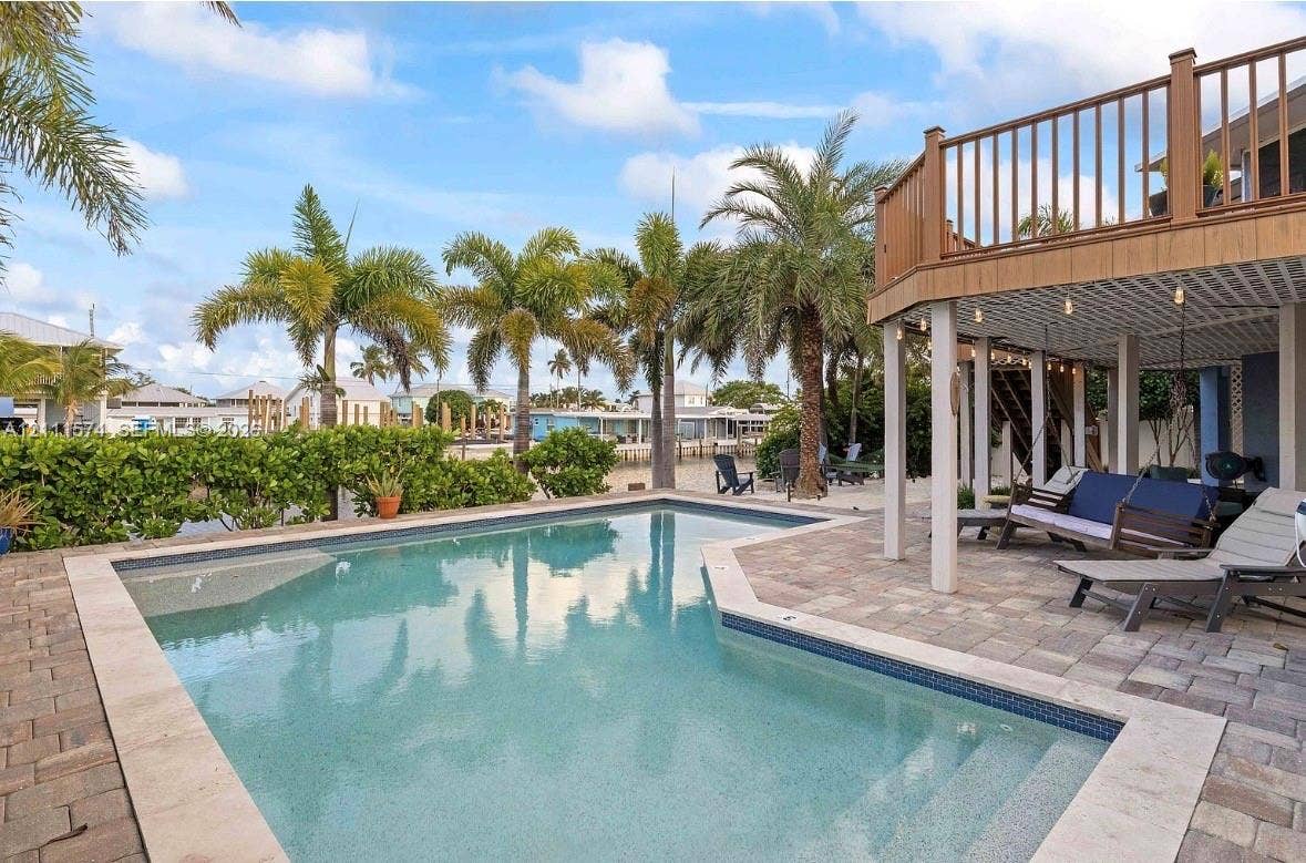 In-ground swimming pool with a stone paver deck, elevated wood-tone balcony, and covered lower-level patio area