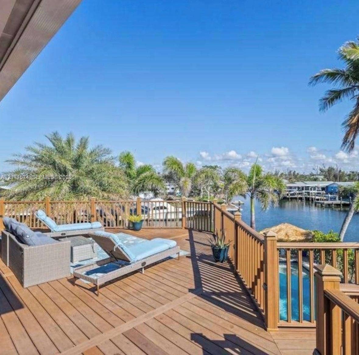 Elevated wooden deck featuring a perimeter railing, overlooking a canal and a swimming pool with mature palm trees