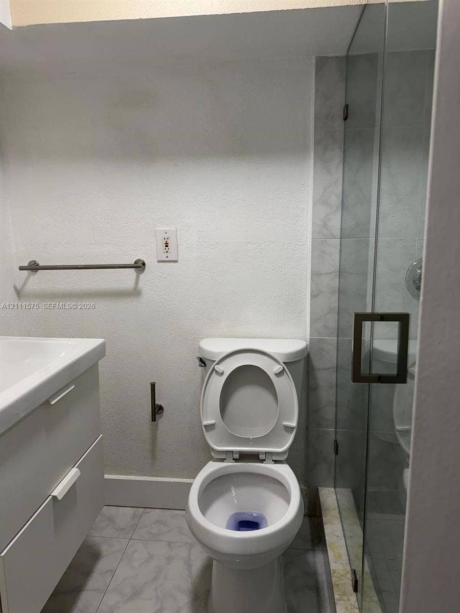 Wall-mounted vanity with integrated basin, floor-to-ceiling glass-enclosed shower, light-toned stone-finish tile flooring, and brushed metal hardware