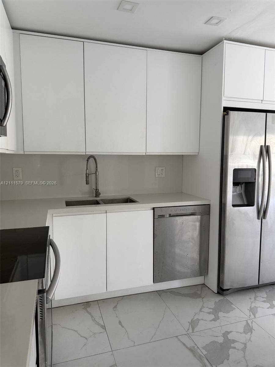 Modern kitchen featuring flat-panel white cabinetry, solid-surface countertops, recessed ceiling lighting, stainless steel appliances, and large-format tile flooring