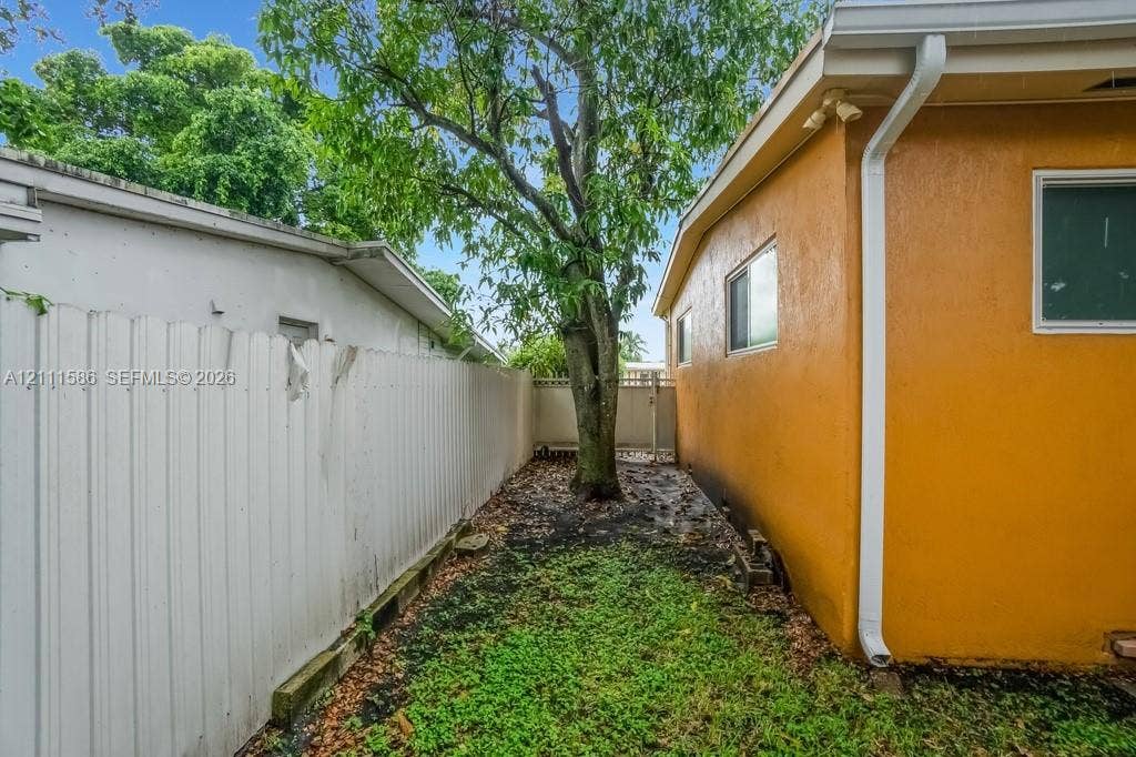 20431 NE 14th Court, Miami, FL 33179 Side yard featuring a stucco exterior, white vinyl privacy fencing, integrated gutter system, and mature tree canopy