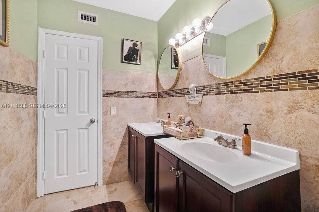 20431 NE 14th Court, Miami, FL 33179 Dual vanity with dark wood-finish cabinetry, integrated white basins, stone-tiled walls with decorative mosaic accent inlay, and circular mirrors with metallic frames