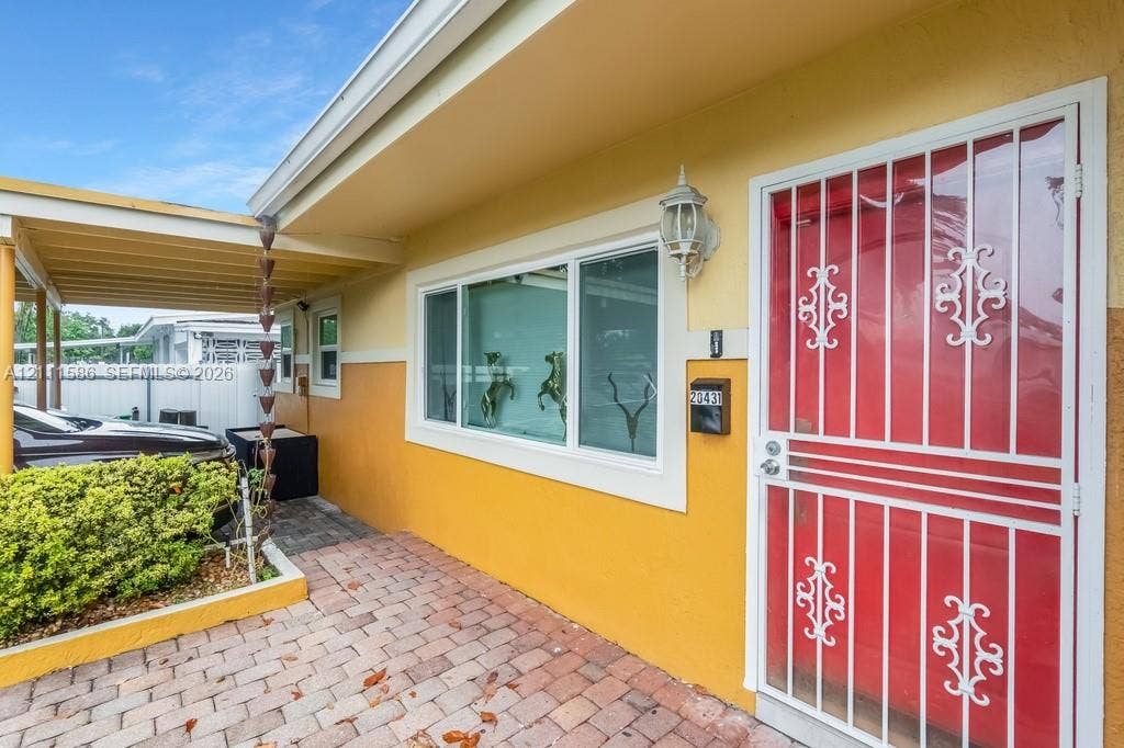 20431 NE 14th Court, Miami, FL 33179 Exterior entryway featuring a paver stone walkway, attached carport, decorative metal security door, wall-mounted exterior light fixture, and chain-style rain gutter
