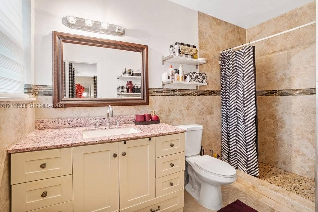 20431 NE 14th Court, Miami, FL 33179 Bathroom featuring a vanity with a speckled countertop, integrated sink, and wood-toned cabinetry, paired with a shower enclosure showcasing natural stone wall tiles, a pebble-finish floor, and a decorative tile inlay