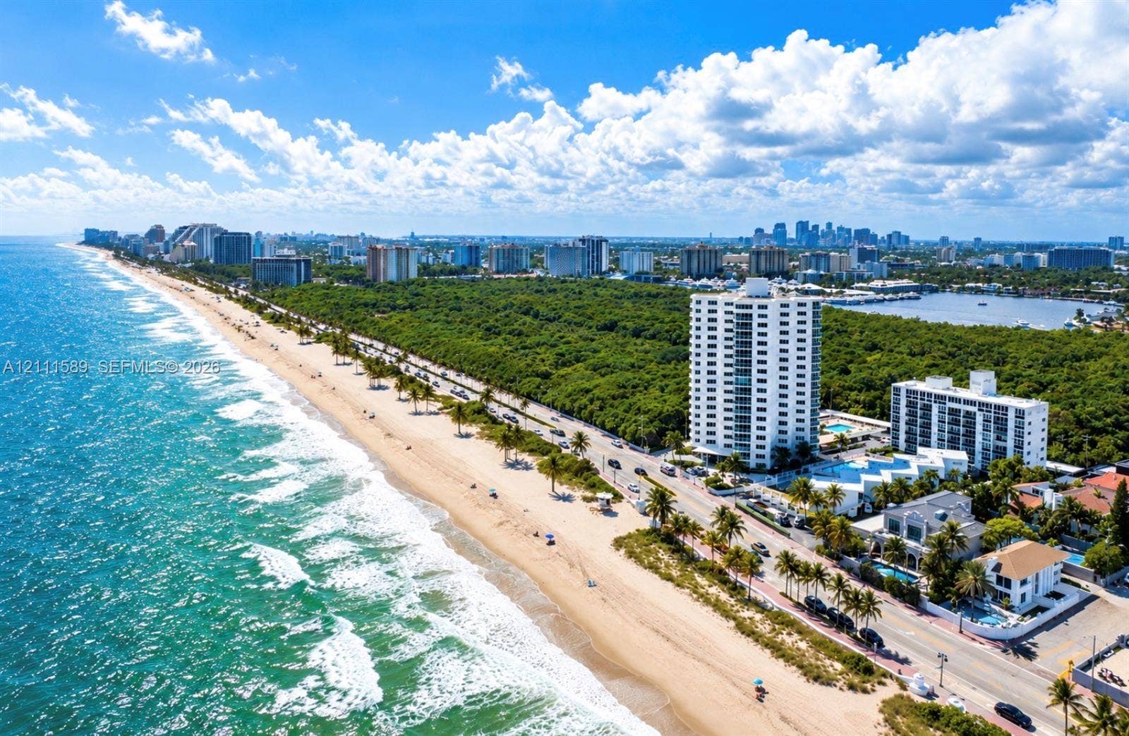 Coastal aerial perspective featuring direct oceanfront access, sandy shoreline, multi-story residential structures, lush tropical vegetation, and distant cityscape