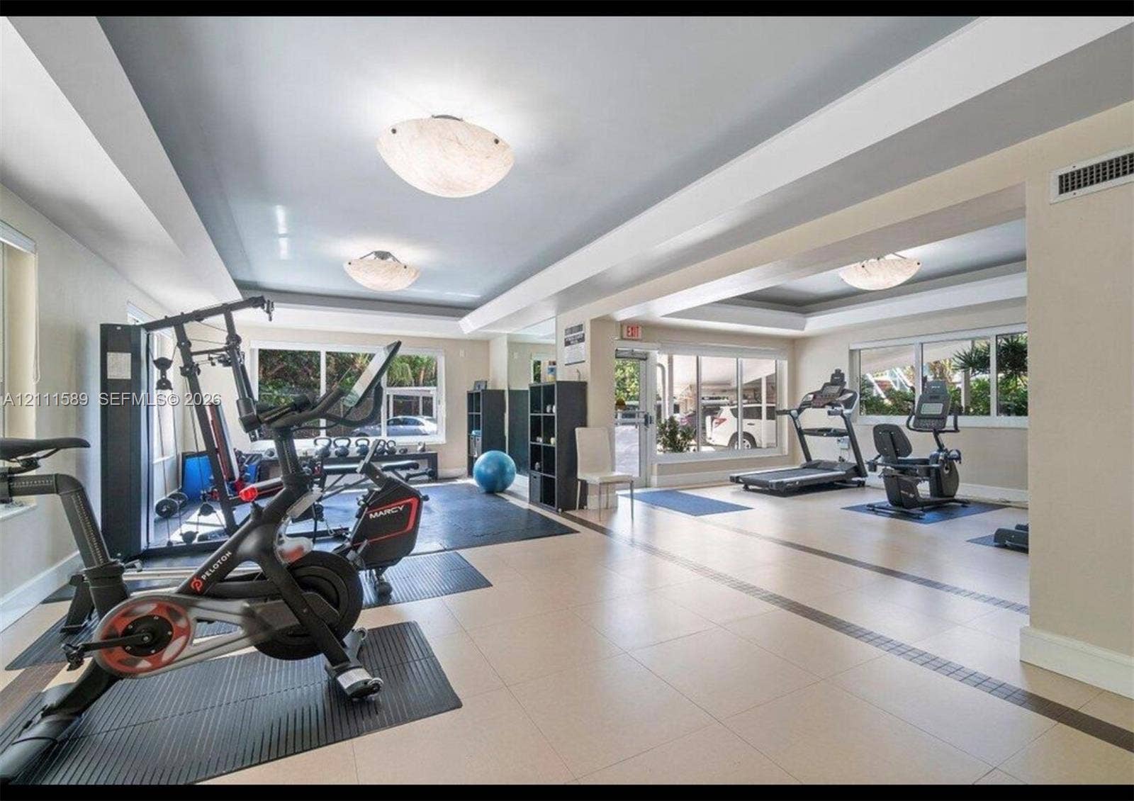 Large fitness room featuring recessed tray ceilings, neutral tile flooring with inlay detail, multiple oversized windows, and semi-flush mount dome lighting