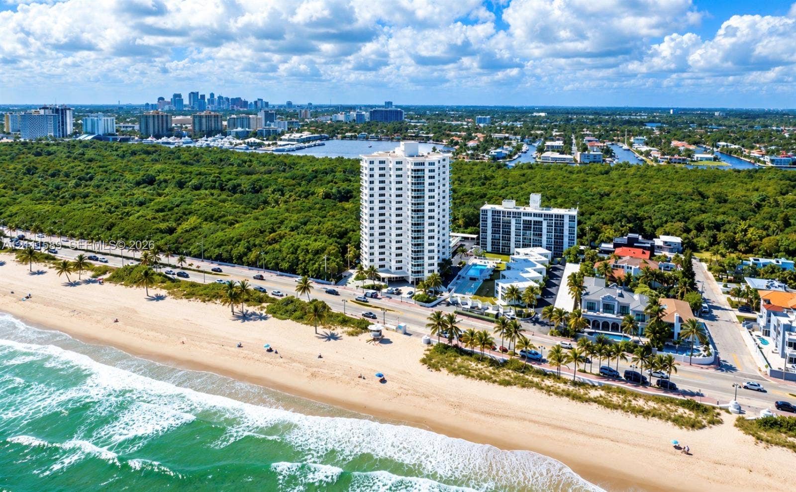 Aerial perspective of a high-rise residential building situated along a coastline featuring direct beachfront access, paved road infrastructure, and proximity to urban skylines