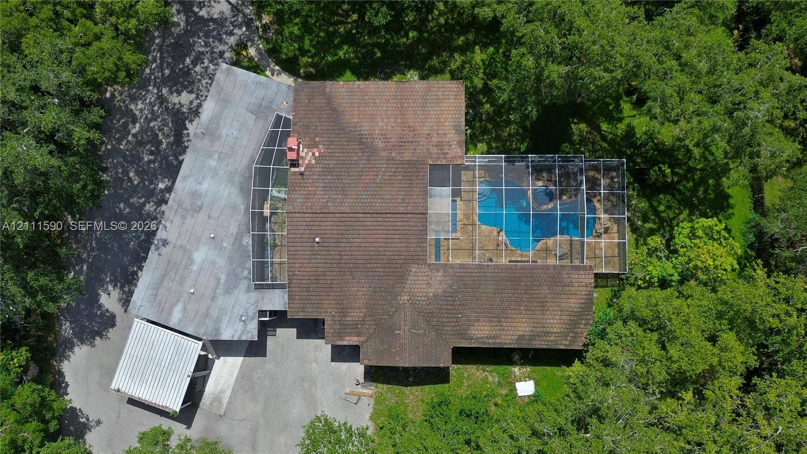 Aerial perspective featuring a multi-level roofline, screened-in swimming pool enclosure, detached covered parking structure, and mature trees on a wooded lot