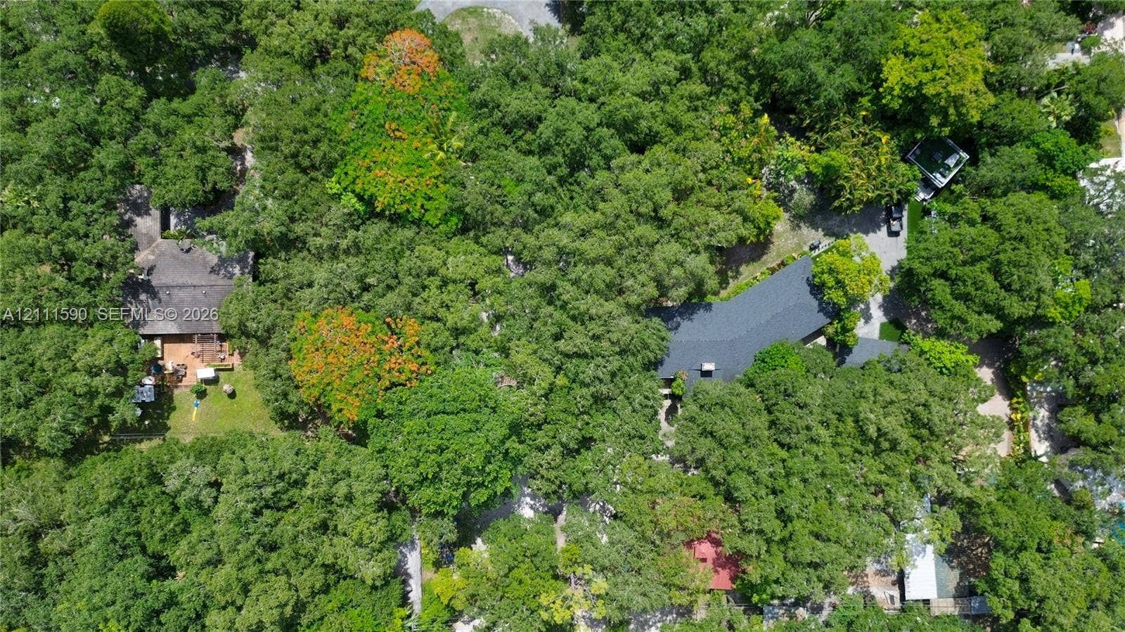 Aerial perspective of a wooded lot featuring mature trees, a multi-section shingled roof, a paved driveway, and established landscape