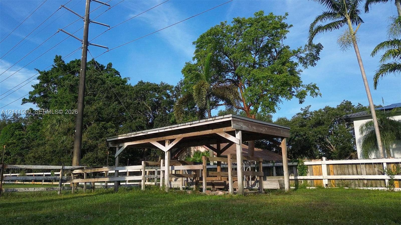 Open-air wood structure featuring a post-and-beam frame, set within a fenced area with mature trees