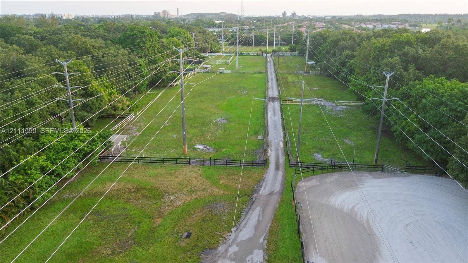 Expansive grounds featuring a central paved driveway, perimeter post-and-rail fencing, a dedicated sand ring, and mature tree buffers