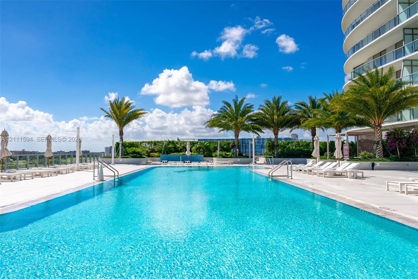 2411 Laguna Circle, #2506, North Miami, FL 33181 Large rectangular swimming pool with metal pool ladders and paved deck, surrounded by mature palm trees and manicured privacy hedging