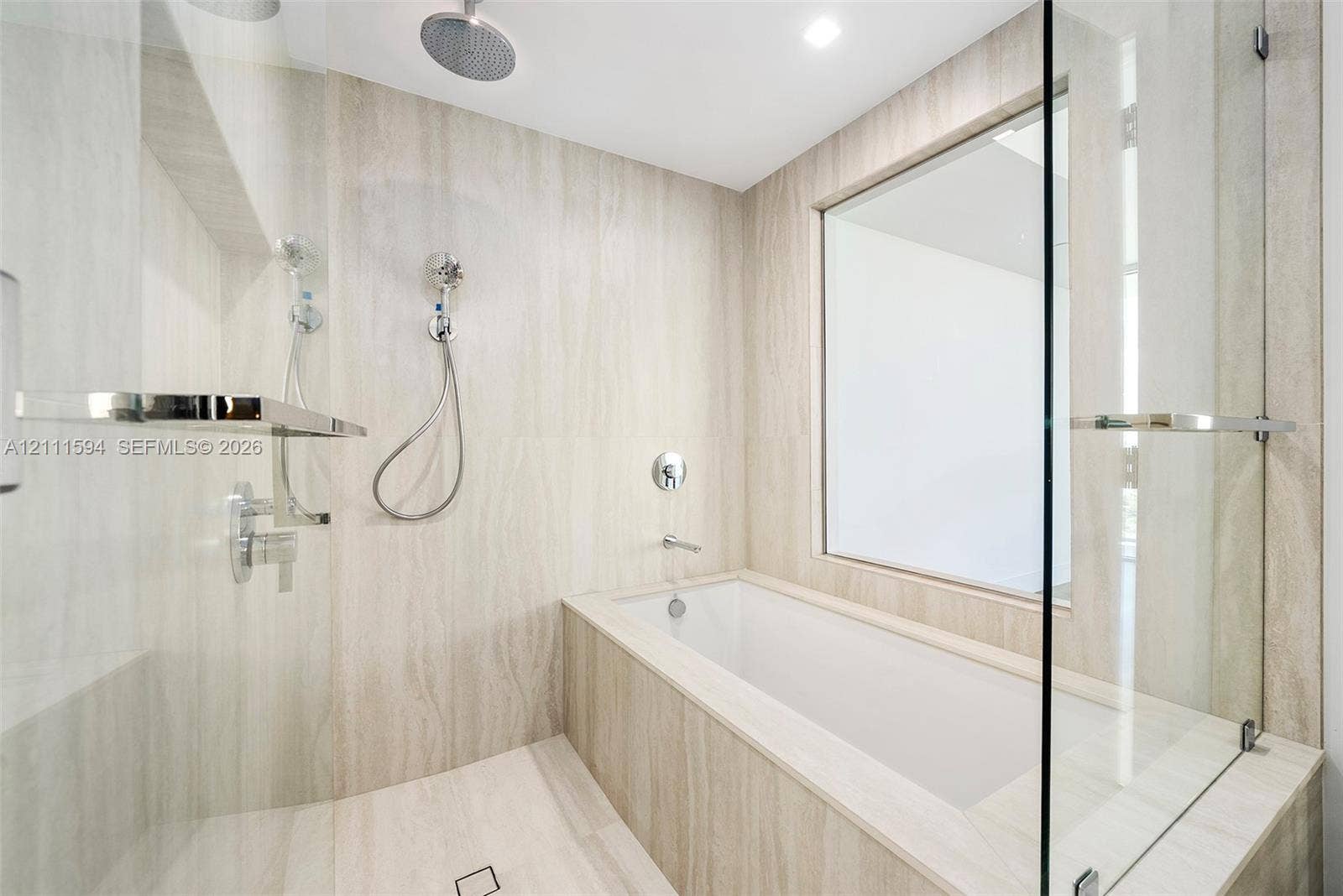 2411 Laguna Circle, #2506, North Miami, FL 33181 Wet room featuring stone-finish wall and floor tiling, deep-soaking bathtub, glass partition, wall-mounted shower system with rain head, and integrated shelving