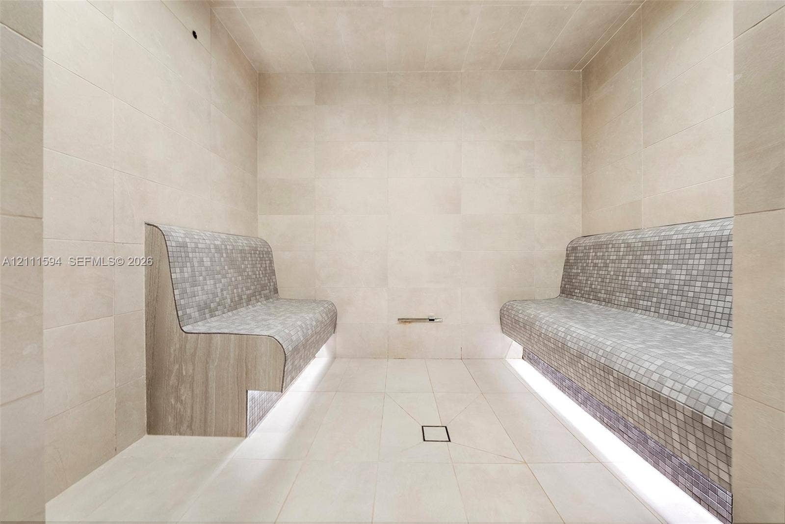 2411 Laguna Circle, #2506, North Miami, FL 33181 Steam room featuring large-format stone wall and floor tile, built-in dual benches with mosaic tile surfacing, integrated under-bench lighting, and a centered floor drain