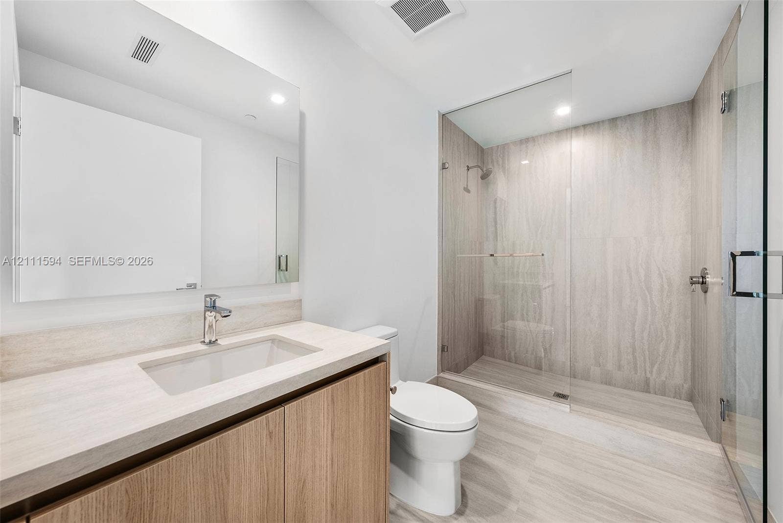 2411 Laguna Circle, #2506, North Miami, FL 33181 Bathroom featuring a wood-finish vanity with an integrated countertop, a frameless glass-enclosed walk-in shower with floor-to-ceiling stone-style tile, and a wall-mounted mirror