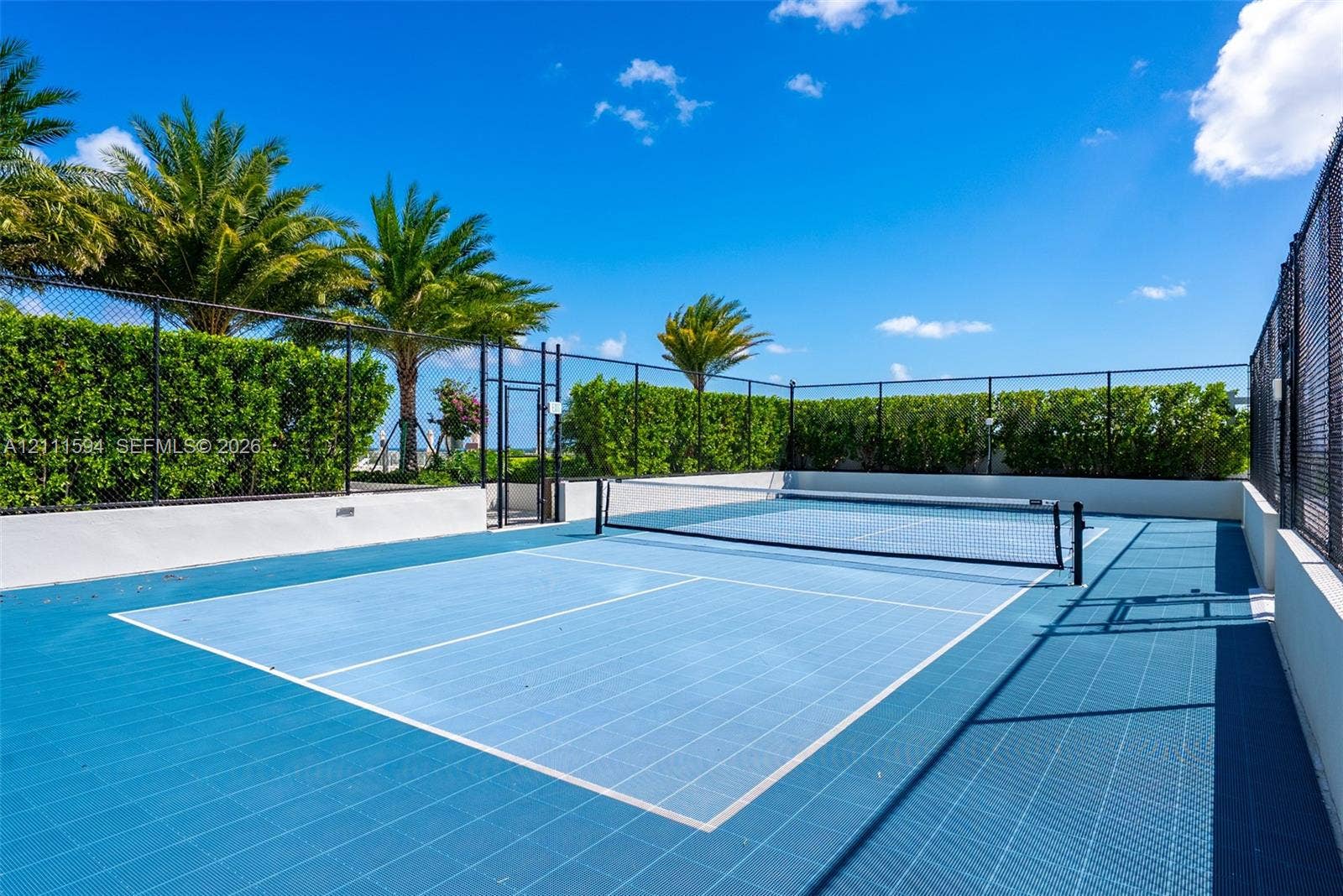 2411 Laguna Circle, #2506, North Miami, FL 33181 Private pickleball court featuring a blue modular surface, perimeter chain-link fencing, and manicured hedge privacy screening