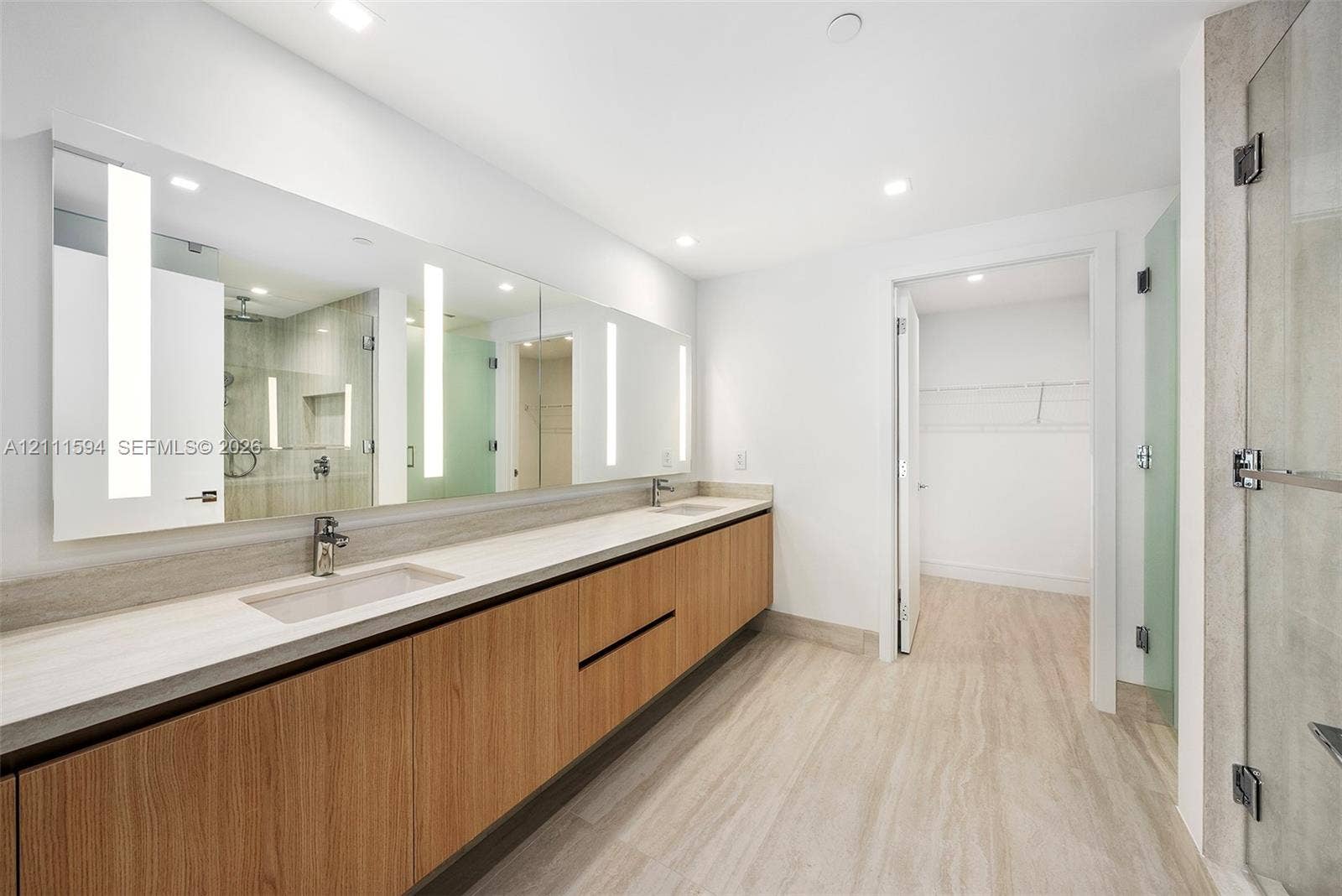 2411 Laguna Circle, #2506, North Miami, FL 33181 Floating wood-finish vanity with dual integrated sinks, expansive wall-to-wall mirror with integrated LED lighting, stone-finish flooring, and glass-enclosed shower